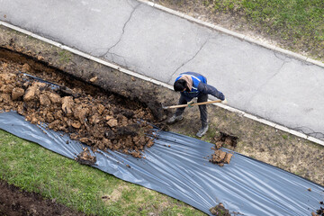 A worker digging a ditch along a pavement in summer.