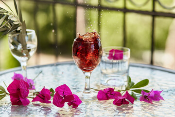 Elegant and colorful explosion of red wine in the glass. Summer bright cocktails on the background. Decorative pink flowers and a branch of an olive tree in a glass. Romantic creative concept.