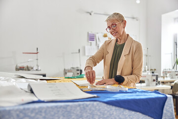 Mature woman designer draws pattern on color fabric at large cutting table in workshop