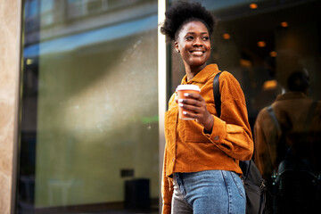 Beautiful young woman using the phone while walking around town. Young african woman drinking coffee outdoors.