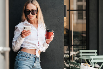 Elegant Woman Standing near the Wall on a Terrace of Cafe, Holding Drink in Hand While Using Smartphone, Confident Female Entrepreneur Wearing Sunglasses Waiting for Meeting at Coffee Shop