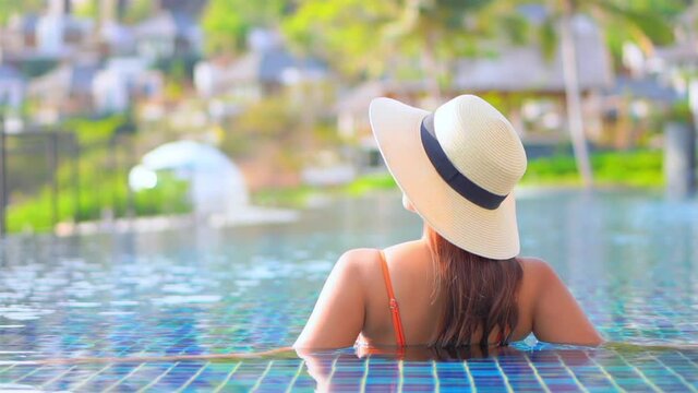 Exotic woman with summer hat enjoying in infinity pool water with splendid view on small tropical resort city, slow motion full frame