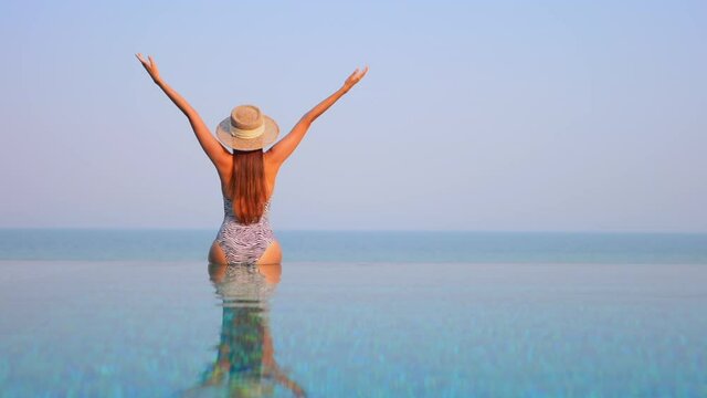 Back of exotic woman sitting on infinity pool border and raising hands, tropical happiness on vacation, slow motion full frame