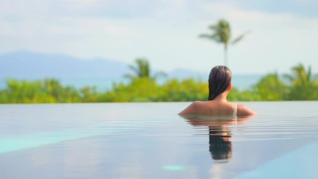 Back of sexy exotic woman in infinity pool enjoying in water, view of a tropical landscape on hot summer day, slow motion full frame