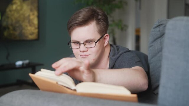 A positive man wearing glasses with down syndrome is reading book lying in the sofa at home