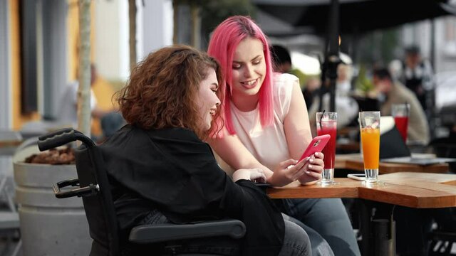 A smiling disabled woman is looking to the smartphone with her friend while sitting on the street cafe outside