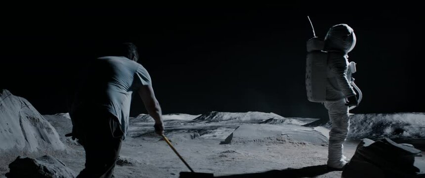 Behind the scenes, actor wearing astronaut suit waiting while film crew decorator straightens lunar surface. Virtual production with LED screens. Shot with 2x anamorphic lens