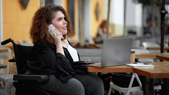 A smiling disabled woman is using her laptop while talking on her smartphone sitting on the street cafe outside