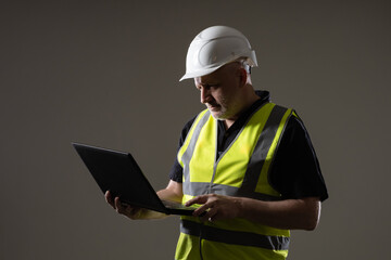 Fototapeta premium Man in construction worker uniform. Human in yellow reflective vest and safety helmet. Portrait of man builder on dark background. Concept engineer uses internet. Builder orders something on Internet