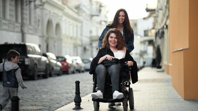 A smiling disabled woman is sitting on the wheelchair while her friend is walking and wheel her wheelchair on the city street
