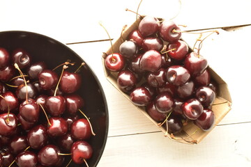 Several ripe organic, dark red cherries in a paper and ceramic plate on a wooden table, close-up, top view.