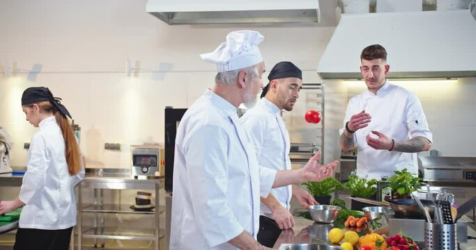 Caucasian male young famous chef speaking with cooks apprentices during working process at restaurant kitchen telling teaching them cooking. Team of chefs preparing food dish. Culinary concept