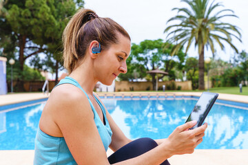 Young fitness woman choosing an online sport class outdoors. Horizontal side view of young woman connecting with technology to a gym class with friends next to a swimming pool.