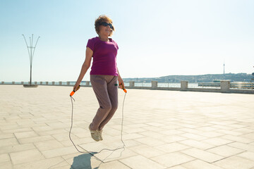 Senior woman with skipping rope doing practice in a public park in the center of town. Healthy lifestyle after retirement. - obrazy, fototapety, plakaty