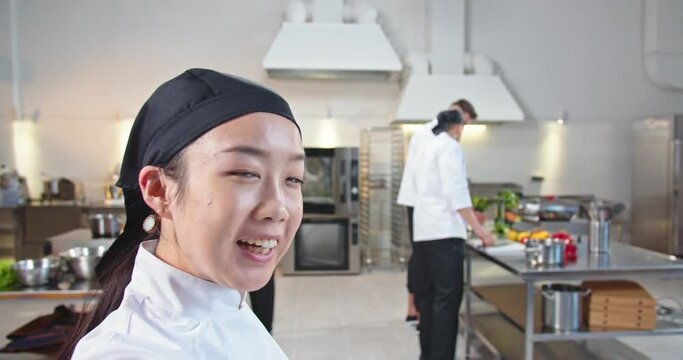 Close up of Asian happy professional female chef videochatting standing at kitchen showing her team of cooks during work process POV of cheerful young woman speaking on video call at restaurant online