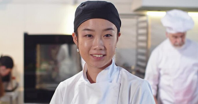Close up portrait of cheerful young Asian beautiful female chef in hat looking at camera in good mood smiling while standing at work at restaurant kitchen. Workflow on background, cooking concept