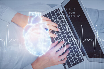 Double exposure of woman hands working on computer and heart hologram drawing. Top View. Medical Science concept.