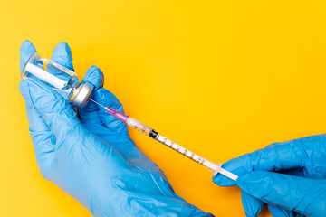 Hands in blue gloves holding syringe and stabbing to a bottle of a vaccine on orange background...