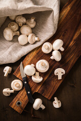 Fototapeta na wymiar Fresh raw white Champignon mushrooms on dark brown moody wooden cutting board with kitchen knife and linen towel