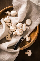 Fresh raw white Champignon mushrooms on linen towel with kitchen knife, wooden bowl and dark brown moody background