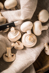 Fresh raw white Champignon mushrooms on linen towel with kitchen knife, wooden bowl and dark brown moody background