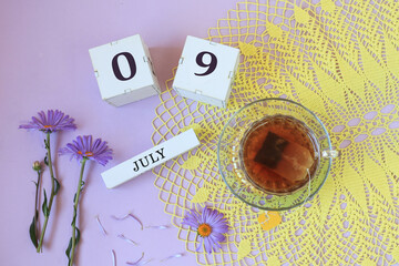 Calendar for July 9: cubes with the numbers 0 and 9, the name of the month of July in English , a cup of tea on a yellow openwork napkin, flowers on a pastel background
