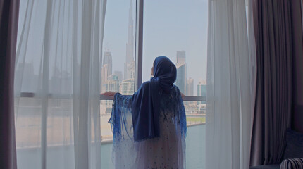 Back view of woman opening curtains and looking out of window to cityscape at hotel in the morning. Modern buildings at the Dubai city