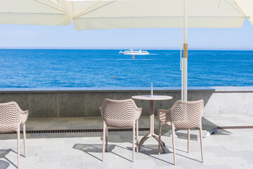 Cafe without visitors on the embankment with a view of the sea, with a floating motor ship. Empty tables under white umbrellas