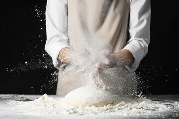 Woman clapping hands and sprinkling flour over dough on dark background