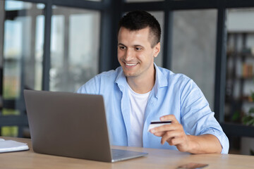 Happy smiling young male or student with a credit card making profitable purchases in the Internet, young entrepreneur or freelancer uses laptop for online shopping sitting at home office