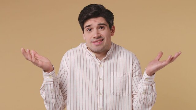 Fun confused shy shamed stylish brunet young man 20s years old wears white striped shirt look camera spread hands say oops ouch oh omg i am so sorry isolated on pastel beige background studio portrait
