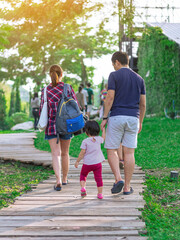 Fototapeta premium Back view of Asian Father hand holding lovely daughter walking on pathway through green garden.Dad and daughter walking together in park. Happy family spending time together outside in green nature.