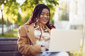 Fototapeta premium African american woman freelancer sitting on a bench in a street and using laptop