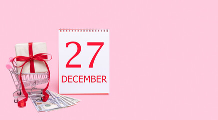 A gift box in a shopping trolley, dollars and a calendar with the date of 27 december on a pink background.