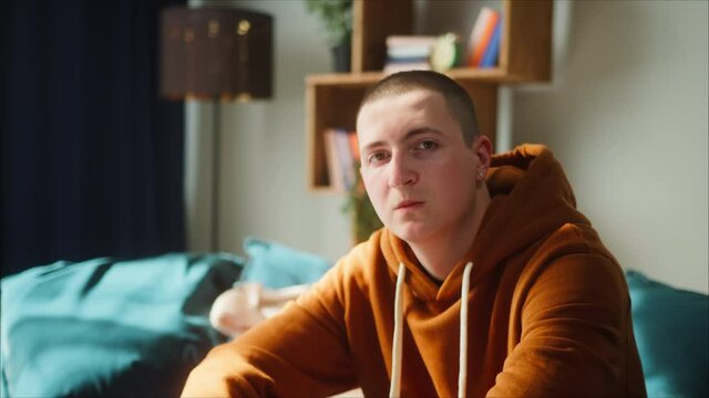 Portrait of brutal teenager with earrings, wearing brown hoodie, young brown-eyed man sitting on sofa and looking in camera with serious face, British model posing. 