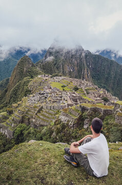 Young man sitting, contemplating Machu Picchu lost city with Huayna Picchu mountain. Ruins of ancient inca civilization in the sacred valley of Cusco Province. Peru, South America