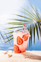 Fresh strawberry beverage with ice, straw in plastic glass on sandy tropical beach with sea view, copy space, vertical. Vacation, relax on coast.