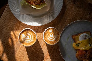 Two coffee cups with latte art in sunlight and breakfast served on wooden table. Eggs benedict with salmon and healthy burger with cappuccino. Morning date in cafe. 