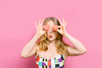 Portrait of a young blonde holding two pink flowers in front of her eyes blowing a kiss with a colourful retro top on a pink background. summer concept. copy space