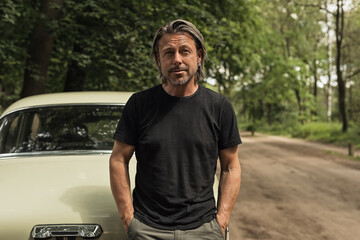 Man in a black t-shirt standing by a classic car on a forest dirt road.