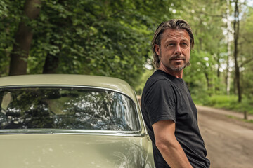 Man in a black t-shirt standing by a classic car on a forest dirt road.