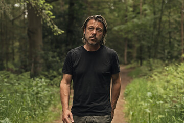Naklejka na ściany i meble Blond man in a black t-shirt walks on a path in a lush forest.