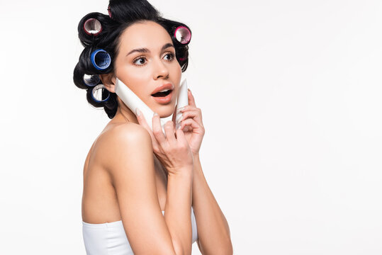 excited young woman with curlers on head holding cream tubes near face with open mouth isolated on white.