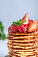 Closeup side view of homemade organic pancakes or crepes decorated with ripe fresh red strawberries and green mint leaves topping served in stack or pile on white wooden table for breakfast. Vertical 