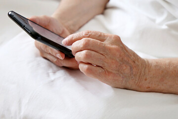 Obraz na płótnie Canvas Elderly woman with smartphone in a bed, mobile phone in wrinkled female hands close up. Concept of online communication in retirement, sms, social media