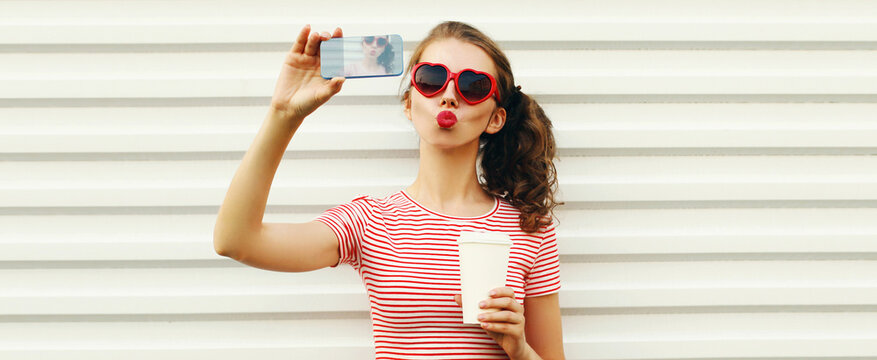 Portrait young woman taking a selfie by smartphone with coffee cup blowing her lips with red lipstick sending sweet air kiss wearing heart shaped sunglasses on white background