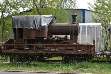 Discarded rusty freight steam locomotive loaded on old wagon standing on blind railroad tracks in abandoned industrial zone. There is grass and trees growing everywhere.