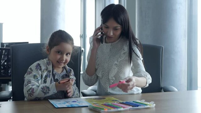Working mom works in the office. Young woman speaks on the phone and cute child in the office draws. Women's business, kindness, care