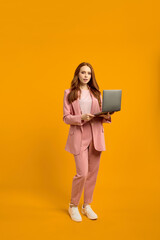 Confident ginger redhaired lady walking carrying laptop isolated on bright yellow orange studio background, copy space. caucasian woman working on laptop, freelance