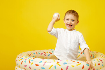 A small smiling boy plays in a dry pool on a yellow background. A child throws a white plastic ball